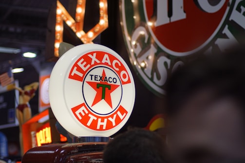 A glowing vintage sign with a circular design displaying the Texaco logo in red, white, and green. It is surrounded by other retro gasoline and oil signs, creating a nostalgic and classic atmosphere.