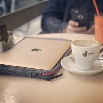 A cozy cafe setting with a closed laptop placed inside a grey fabric case on a creamy white table. Beside it, a white cup of coffee with some remaining froth sits on a saucer, branded with 'danesi caffe'. In the blurred background, a person is holding a smartphone, and there is a tall glass filled with an orange beverage.