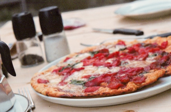 A pizza topped with cheese, tomato, and herbs is served on a white plate. In the background, there are salt and pepper shakers, a fork, and a butter knife placed on a wooden table, suggesting a casual dining setting.