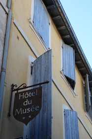 A rustic building facade in a European style features a sign with 'Hôtel du Musée' written on it. The walls are painted in a soft yellow, accented by pale blue wooden shutters that cover the windows. The scene suggests an old-world charm, with a simple yet elegant aesthetic.