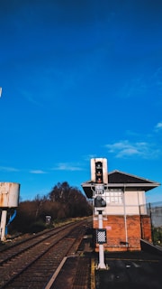 A railway track leading through an area with a signal and a small building nearby. The sky is clear with a vibrant blue color, and there are trees and vegetation on either side of the tracks. The structure next to the track has a brick base and a white upper section.
