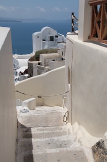 A narrow stone staircase leads downwards, surrounded by whitewashed walls typical of Greek island architecture. In the background, the deep blue sea meets the sky, and traditional buildings with rounded roofs and small windows are visible. Some greenery is incorporated into the architecture, and the overall scene captures a serene coastal setting.