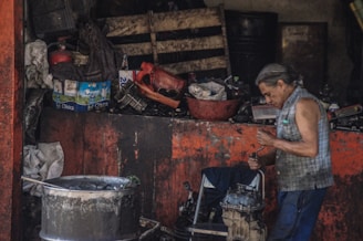 A man with gray hair is engaged in working on a mechanical component in a cluttered workshop. The surroundings are filled with various tools, parts, and materials such as a large metallic barrel, a red container, cardboard boxes, and other equipment.