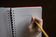 A hand holding a yellow pencil is writing 'Thought Catalog' on a lined notebook. The notebook has a red cover and is spiral-bound, resting on a dark surface.
