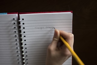 A hand holding a yellow pencil is writing 'Thought Catalog' on a lined notebook. The notebook has a red cover and is spiral-bound, resting on a dark surface.