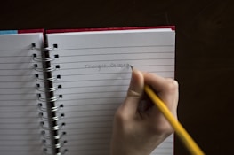 A hand holding a yellow pencil is writing 'Thought Catalog' on a lined notebook. The notebook has a red cover and is spiral-bound, resting on a dark surface.