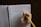 A hand holding a yellow pencil is writing 'Thought Catalog' on a lined notebook. The notebook has a red cover and is spiral-bound, resting on a dark surface.