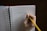 A hand holding a yellow pencil is writing 'Thought Catalog' on a lined notebook. The notebook has a red cover and is spiral-bound, resting on a dark surface.