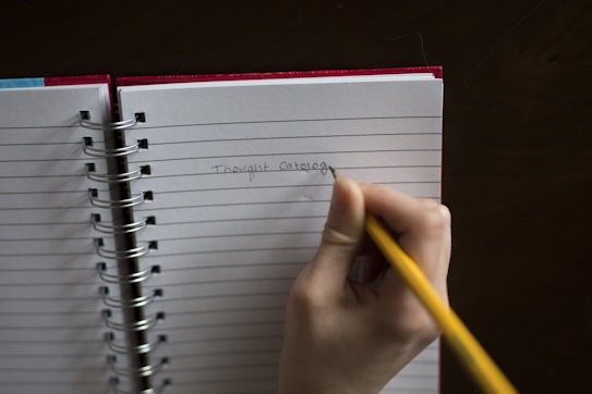 A hand holding a yellow pencil is writing 'Thought Catalog' on a lined notebook. The notebook has a red cover and is spiral-bound, resting on a dark surface.