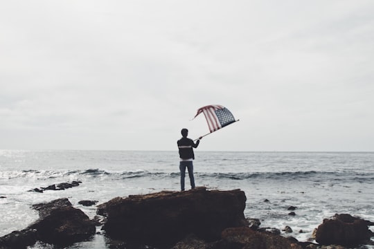 man holding flag of America standing on boulder near seashore in Rancho Palos Verdes United States