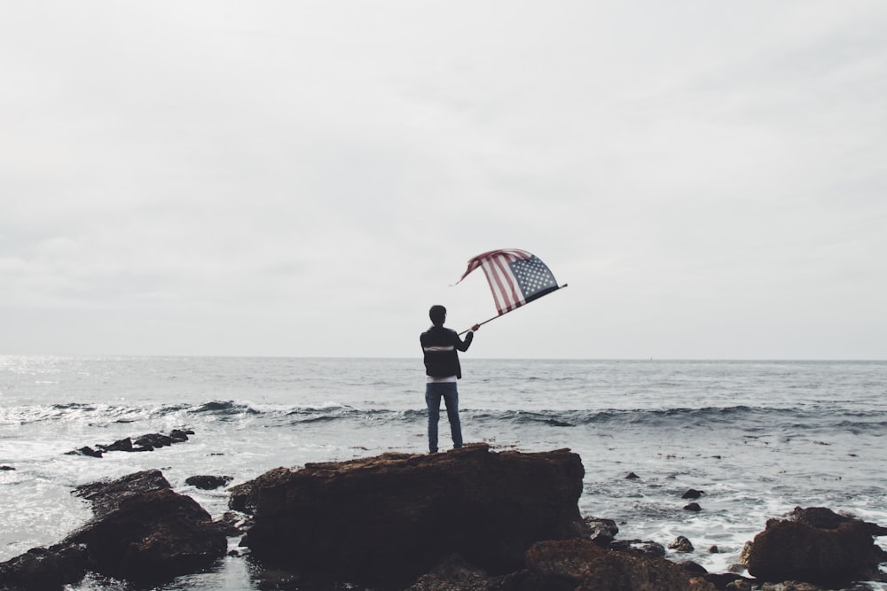 Mann mit Amerika-Flagge steht auf Felsbrocken in der Nähe der Küste