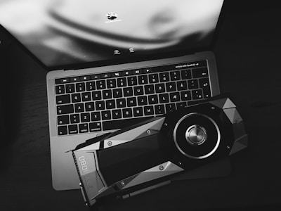 A black and white image displaying a laptop with an illuminated keyboard and a large graphics card placed on top of it. The screen shows a blurred image and login options.