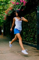 A woman in motion, possibly jogging or dancing, wears a white tank top and blue shorts. Her white sneakers step lightly on a paved path bordered by a tall fence covered in green foliage and vibrant pink flowers. Her hair flows freely, adding to the dynamic sense of movement.