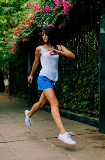 A woman in motion, possibly jogging or dancing, wears a white tank top and blue shorts. Her white sneakers step lightly on a paved path bordered by a tall fence covered in green foliage and vibrant pink flowers. Her hair flows freely, adding to the dynamic sense of movement.