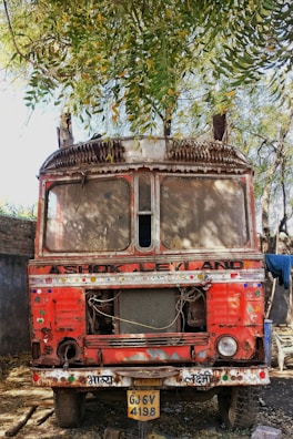 An old, rusted truck with a red body and visible wear and tear is parked under a large tree. The front of the truck shows significant damage and missing parts, such as headlights and the bumper. Bright patches of paint and decorative stickers adorn the truck's surface. A license plate with the number GJ 6V 4198 is attached to the front. The scene is shaded by the dense foliage of the tree above.