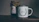 A rustic setting features a jar of organic certified honey with a handmade paper cover next to a white enamel mug. The mug is adorned with an illustration of green leaves encircling the words 'Head Gardener.' These items are placed on a wooden surface, suggesting a cozy, natural atmosphere.
