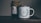 A rustic setting features a jar of organic certified honey with a handmade paper cover next to a white enamel mug. The mug is adorned with an illustration of green leaves encircling the words 'Head Gardener.' These items are placed on a wooden surface, suggesting a cozy, natural atmosphere.