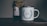 A rustic setting features a jar of organic certified honey with a handmade paper cover next to a white enamel mug. The mug is adorned with an illustration of green leaves encircling the words 'Head Gardener.' These items are placed on a wooden surface, suggesting a cozy, natural atmosphere.
