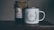 A rustic setting features a jar of organic certified honey with a handmade paper cover next to a white enamel mug. The mug is adorned with an illustration of green leaves encircling the words 'Head Gardener.' These items are placed on a wooden surface, suggesting a cozy, natural atmosphere.
