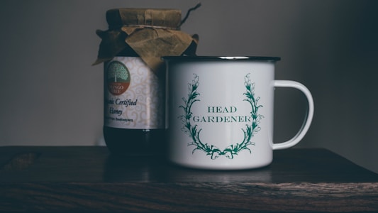 A rustic setting features a jar of organic certified honey with a handmade paper cover next to a white enamel mug. The mug is adorned with an illustration of green leaves encircling the words 'Head Gardener.' These items are placed on a wooden surface, suggesting a cozy, natural atmosphere.
