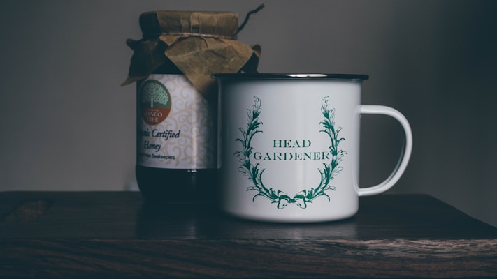A rustic setting features a jar of organic certified honey with a handmade paper cover next to a white enamel mug. The mug is adorned with an illustration of green leaves encircling the words 'Head Gardener.' These items are placed on a wooden surface, suggesting a cozy, natural atmosphere.