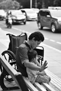 A black and white image depicting a man sitting on a bench beside a wheelchair while holding a child in his lap. The background features a busy street with cars and a motorcycle passing by.