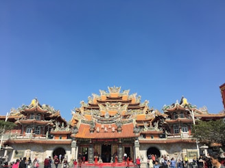 An intricately designed Asian temple featuring ornate, colorful roofs with detailed carvings. The temple is bustling with people walking and taking pictures, set against a bright blue sky.