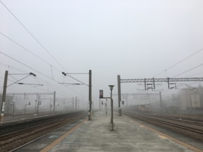 A deserted train station platform enveloped in heavy fog. Overhead power lines extend into the distance, with multiple tracks visible on either side. A solitary lamp post stands in the middle of the platform, and signs with numbers are visible, denoting the tracks.