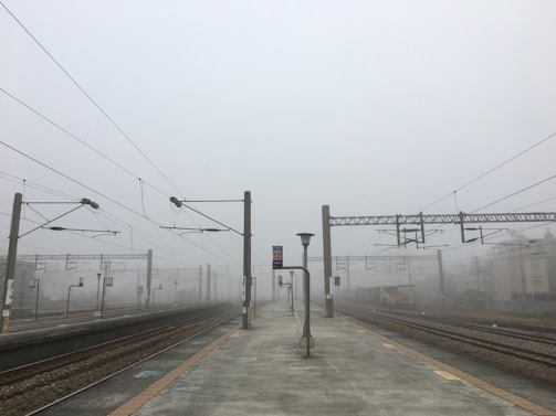 A deserted train station platform enveloped in heavy fog. Overhead power lines extend into the distance, with multiple tracks visible on either side. A solitary lamp post stands in the middle of the platform, and signs with numbers are visible, denoting the tracks.