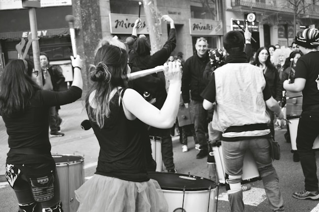 A street scene featuring a group of people engaging in a drum performance. The participants are holding drumsticks and appear animated and focused, surrounded by a diverse crowd of onlookers. The atmosphere seems lively and communal.