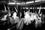A group of people is dressed in long robes with tall, pointed hoods. They stand in a room that resembles a church or other large hall, with benches on either side. The black-and-white photograph highlights the contrast between the dark hoods and lighter garments, creating a dramatic effect.