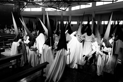 A group of people is dressed in long robes with tall, pointed hoods. They stand in a room that resembles a church or other large hall, with benches on either side. The black-and-white photograph highlights the contrast between the dark hoods and lighter garments, creating a dramatic effect.
