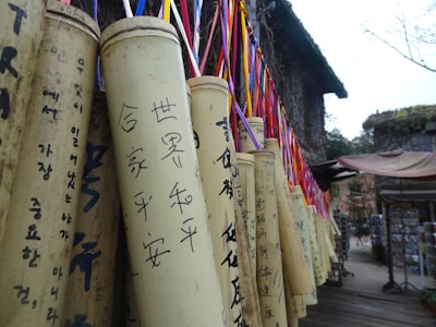 A series of bamboo tubes hang vertically, each intricately inscribed with different calligraphic writing. Colorful ribbons are attached to the tops of the tubes, adding a vibrant touch to the otherwise muted tones. The scene appears to be in an outdoor area with traditional architecture visible in the background, including a structure with a moss-covered roof and a wooden platform.