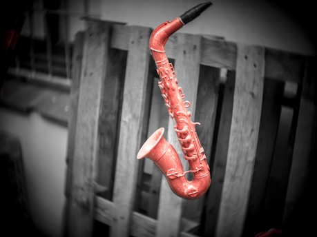 A red saxophone prominently stands out against a monochrome background. The instrument appears suspended or potentially floating in front of a wooden pallet.