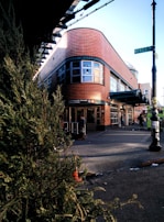 A modern building with a curved brick facade and large windows under a clear blue sky. A street corner with a visible sign reading 'Myrtle Ave' and a subway entrance is present in the foreground. People are seen walking near the entrance, and a leafy green bush partially obscures the left side of the image.