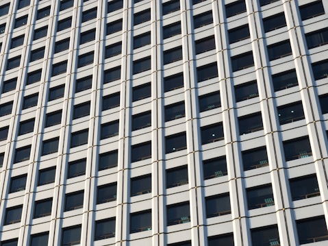 The image features the facade of a modern high-rise building with a grid pattern of windows. The structure's design relies heavily on vertical and horizontal lines, creating a repetitive geometric pattern. The window panels are dark, contrasting with the lighter framework, giving it a sleek and organized appearance.