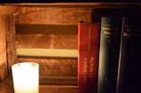A wooden bookshelf displays three hardcover books standing vertically, illuminated by the warm light of a candle placed nearby. The candle is encased in a glass holder, casting a soft glow on the surroundings. The visible book titles include works by Miguel de Cervantes, Arthur Conan Doyle, and Charles Baudelaire.
