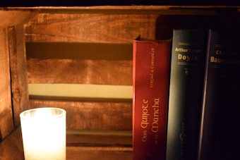 A wooden bookshelf displays three hardcover books standing vertically, illuminated by the warm light of a candle placed nearby. The candle is encased in a glass holder, casting a soft glow on the surroundings. The visible book titles include works by Miguel de Cervantes, Arthur Conan Doyle, and Charles Baudelaire.