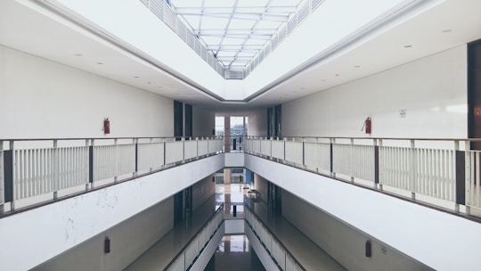 A symmetrical indoor corridor with two levels connected by a balcony running along both sides. The walls are white and the ceiling above is glass, allowing natural light to illuminate the space. Fire extinguishers are mounted at intervals along the walls.