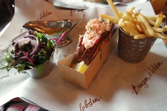 A meal consisting of lobster roll with a toasted bun and lobster filling, accompanied by a metal cup of French fries on the right and a small salad with mixed greens and red onion slices on the left. The dishes are arranged on branded paper.