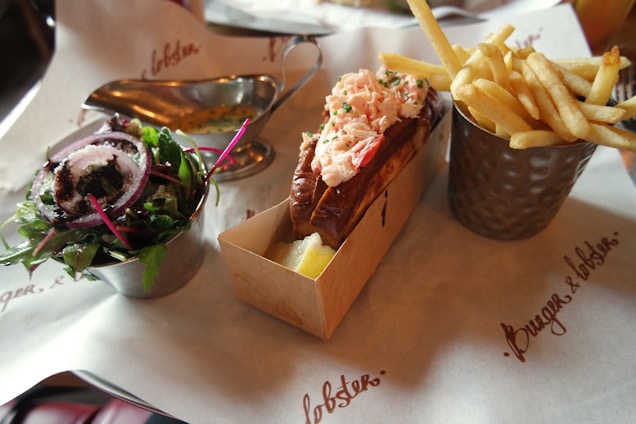 A meal consisting of lobster roll with a toasted bun and lobster filling, accompanied by a metal cup of French fries on the right and a small salad with mixed greens and red onion slices on the left. The dishes are arranged on branded paper.