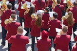 A group of musicians is gathered in formation, each wearing a matching maroon jacket. They are holding brass instruments, such as trumpets and French horns, with musical scores in front of them. The scene appears to be outdoors in a bright setting.