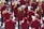A group of musicians is gathered in formation, each wearing a matching maroon jacket. They are holding brass instruments, such as trumpets and French horns, with musical scores in front of them. The scene appears to be outdoors in a bright setting.