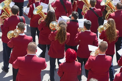 A group of musicians is gathered in formation, each wearing a matching maroon jacket. They are holding brass instruments, such as trumpets and French horns, with musical scores in front of them. The scene appears to be outdoors in a bright setting.
