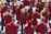 A group of musicians is gathered in formation, each wearing a matching maroon jacket. They are holding brass instruments, such as trumpets and French horns, with musical scores in front of them. The scene appears to be outdoors in a bright setting.