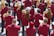 A group of musicians is gathered in formation, each wearing a matching maroon jacket. They are holding brass instruments, such as trumpets and French horns, with musical scores in front of them. The scene appears to be outdoors in a bright setting.