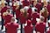 A group of musicians is gathered in formation, each wearing a matching maroon jacket. They are holding brass instruments, such as trumpets and French horns, with musical scores in front of them. The scene appears to be outdoors in a bright setting.