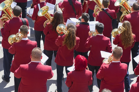 A group of musicians is gathered in formation, each wearing a matching maroon jacket. They are holding brass instruments, such as trumpets and French horns, with musical scores in front of them. The scene appears to be outdoors in a bright setting.