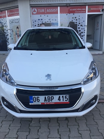 A white car parked in front of a storefront with glass doors featuring signage. The car is positioned on a textured brick pavement, and the storefront has a logo and additional text signage visible. The car's license plate is displayed, and the Peugeot emblem is prominently located on the car's bonnet.
