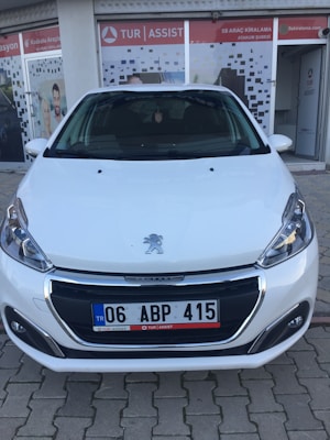 A white car parked in front of a storefront with glass doors featuring signage. The car is positioned on a textured brick pavement, and the storefront has a logo and additional text signage visible. The car's license plate is displayed, and the Peugeot emblem is prominently located on the car's bonnet.